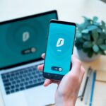 Dłoń trzymająca smartfon z aplikacją VPN, w tle laptop na biurku