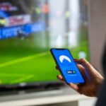 Osoba trzyma smartfon z aplikacją VPN podczas oglądania sportu w TV